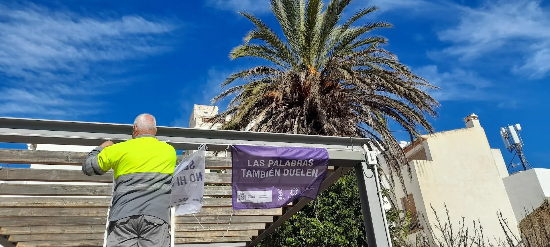A man hanging a purple banner reading “Las palabras también duelen” from a balcony in Benitachell